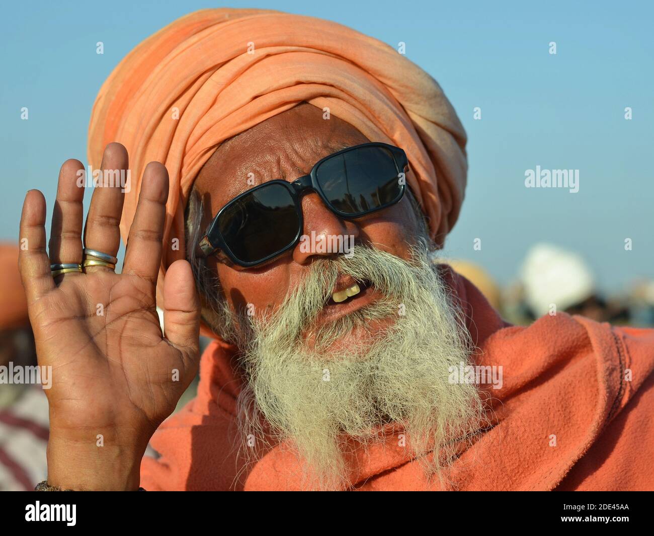 Old Indian Hindu sadhu baba with orange turban and dark sunglasses raises his right hand in order to greet and offer a blessing. Stock Photo