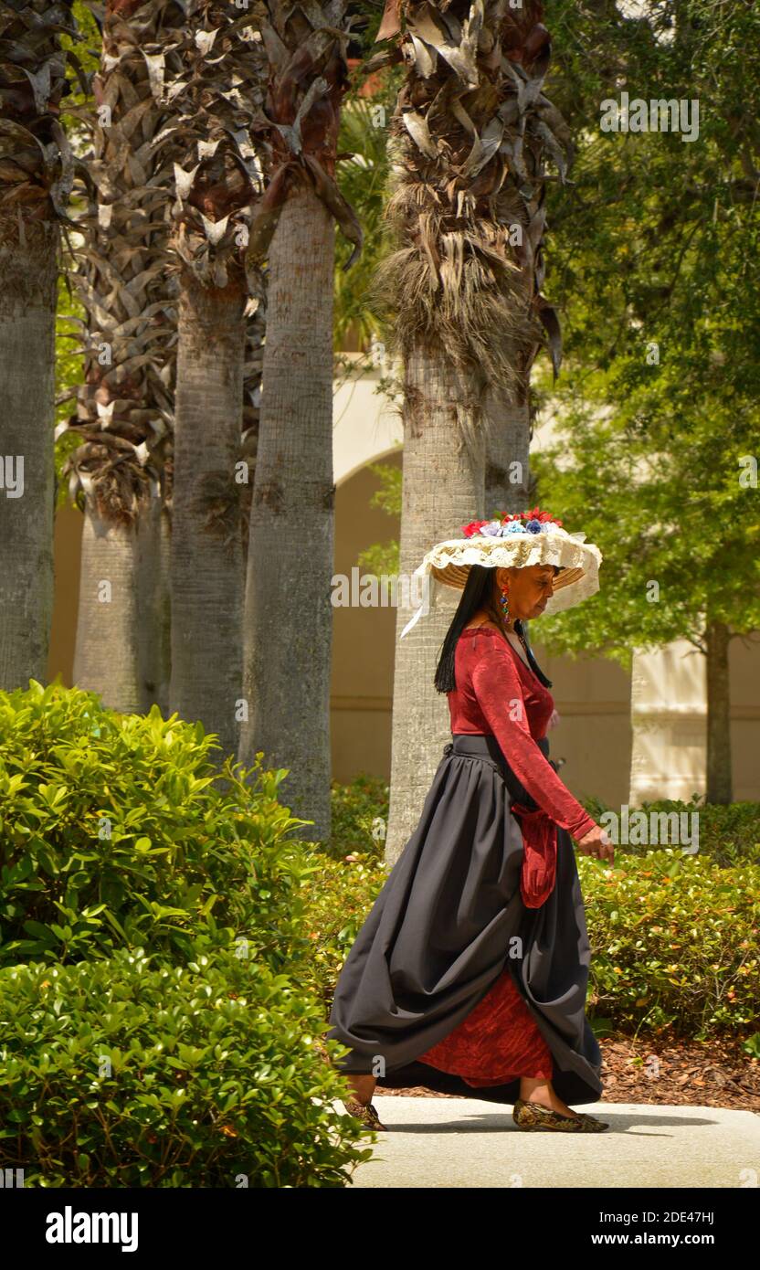 A lovely cultural flash of a woman of color wearing festive Spanish summer hat with lace and flowers and red velvet dress walking in downtown amongst Stock Photo