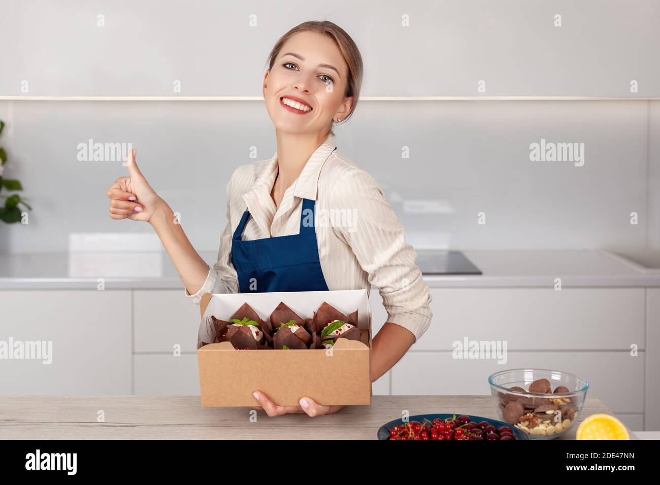 Young smiling woman baker in uniform holding paper delivery packaging box with ready cupcakes or muffins and showing thumb up. Concept of successful f Stock Photo