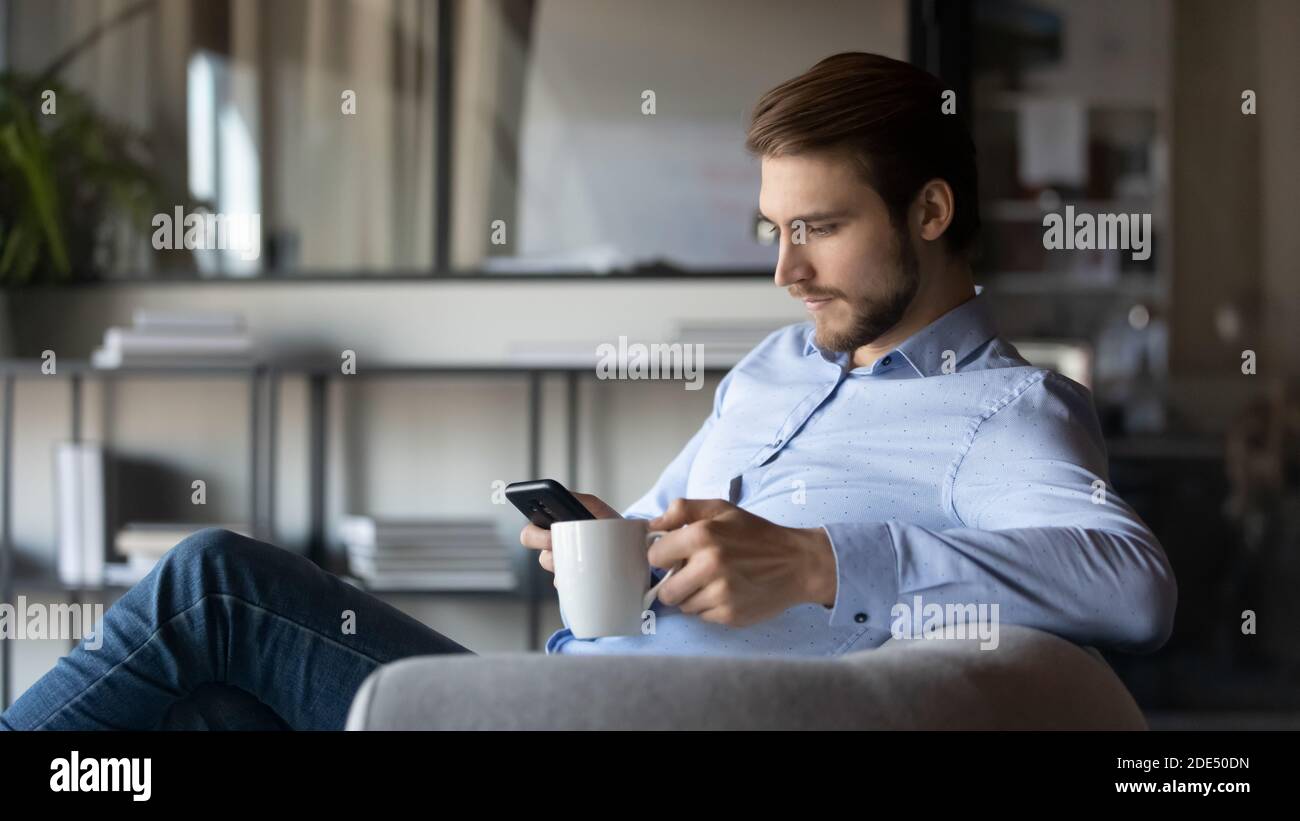 Pensive young businessman focused on smartphone holding cup of tea Stock Photo