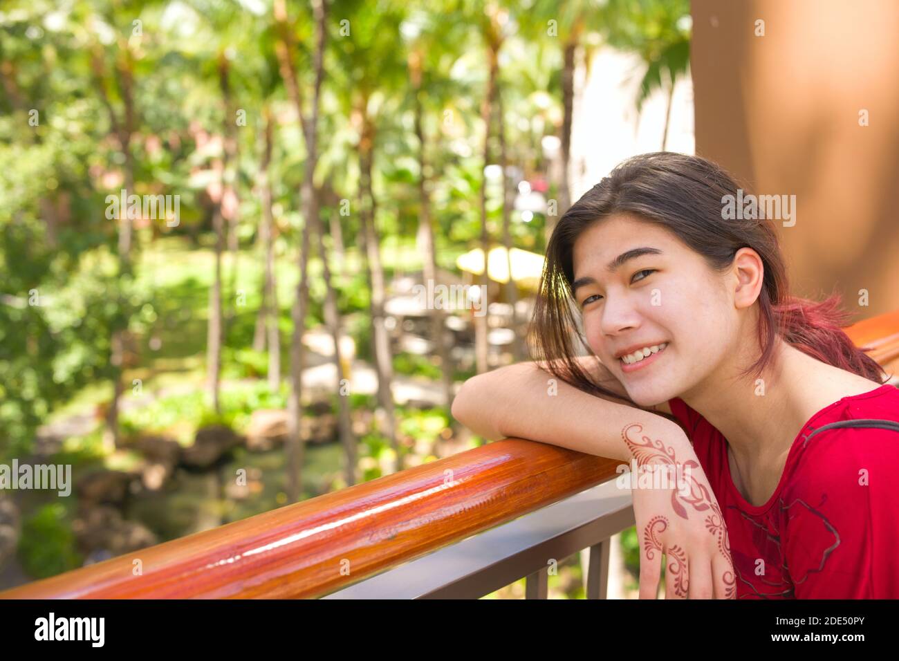 Biracial teen female smiling as she leans on railing overlooking palm and coconut trees on sunny day in Hawaii Stock Photo
