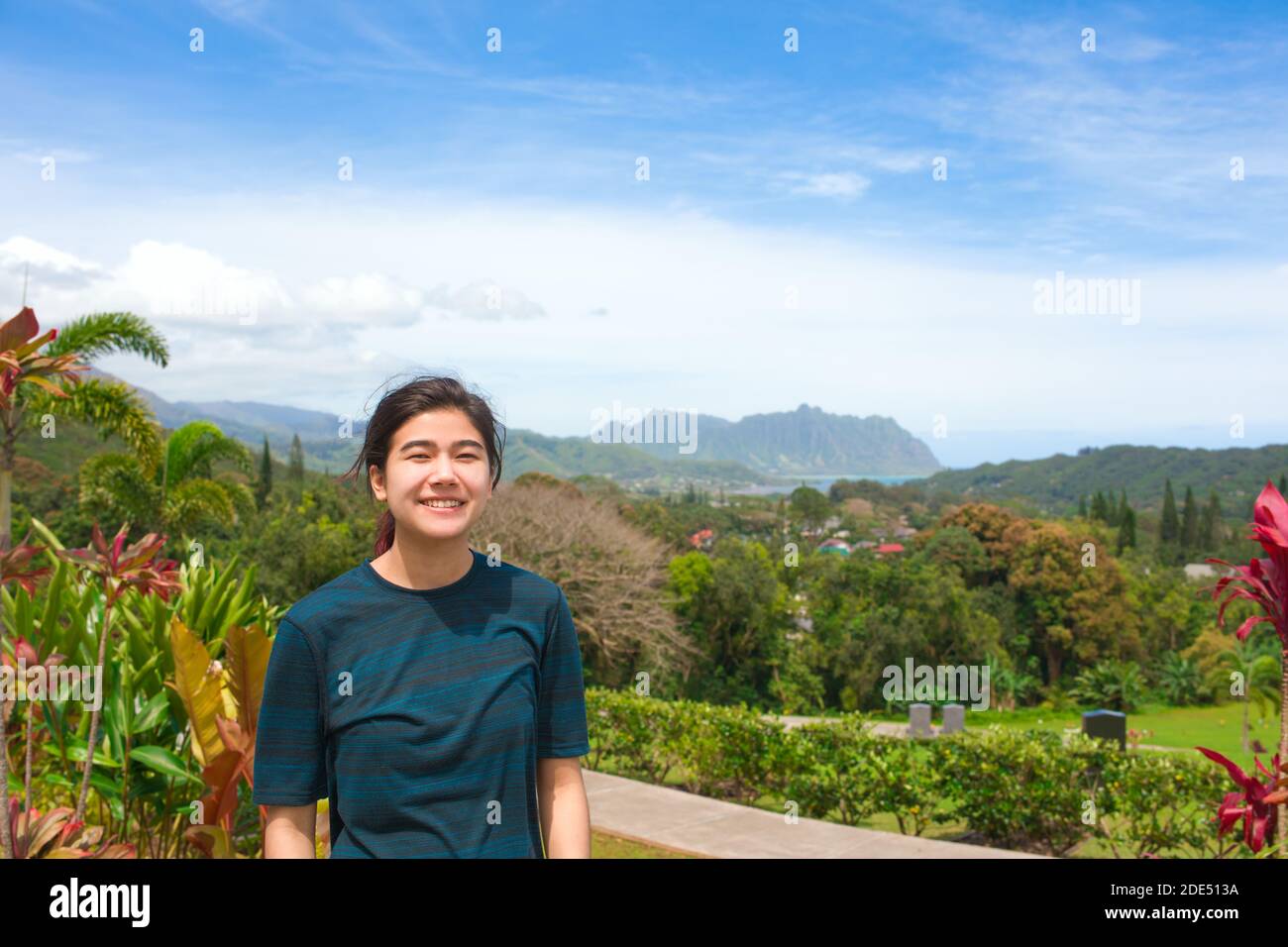 Smiling biracial teen girl outdoors in nature on sunny day with tropical ocean and mountains in background on island of Oahu, Hawaii Stock Photo