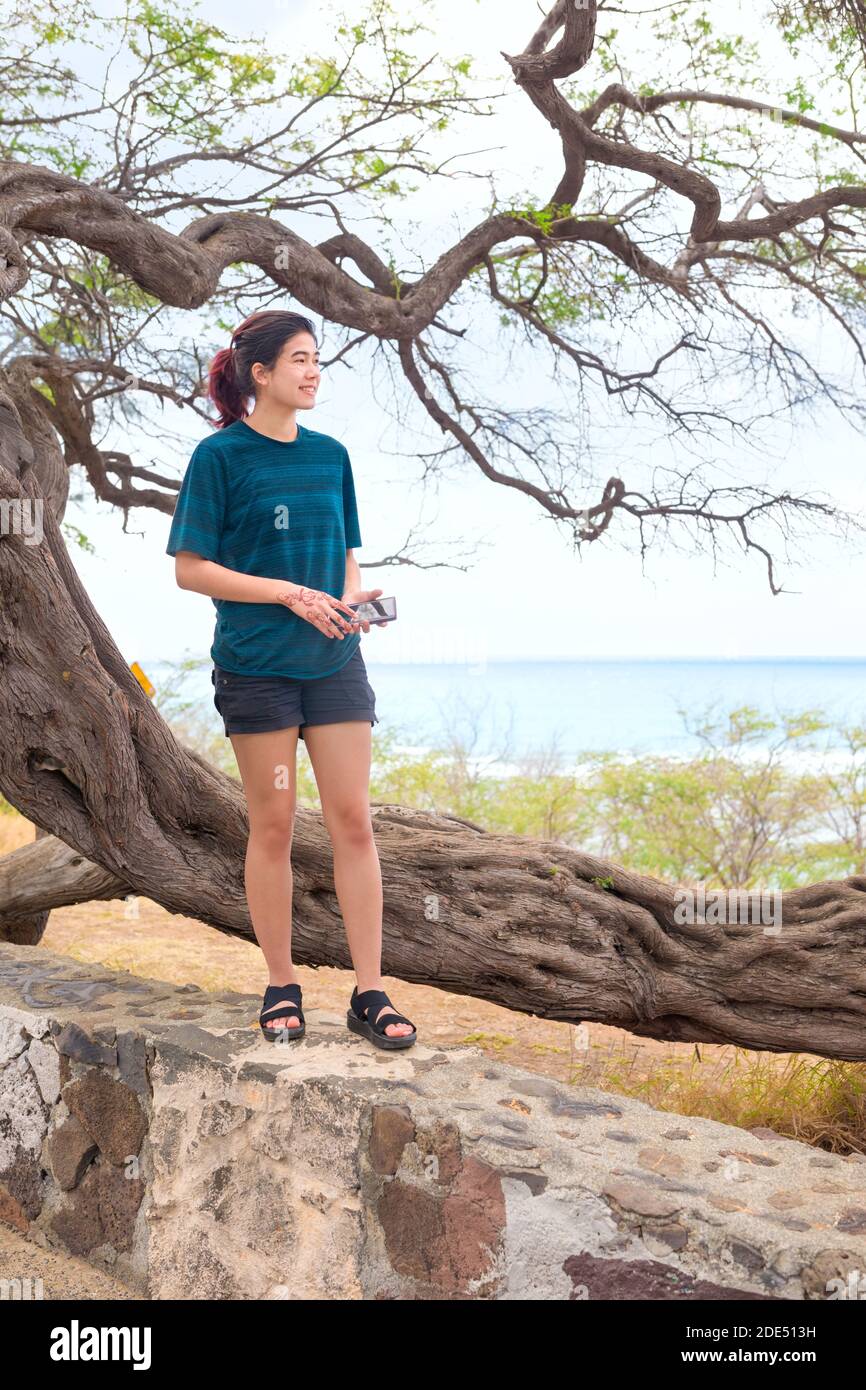 Biracial teen girl standing on stone wall by a gnarled and twisted windswept tree looking out towards blue ocean horizon in background Stock Photo
