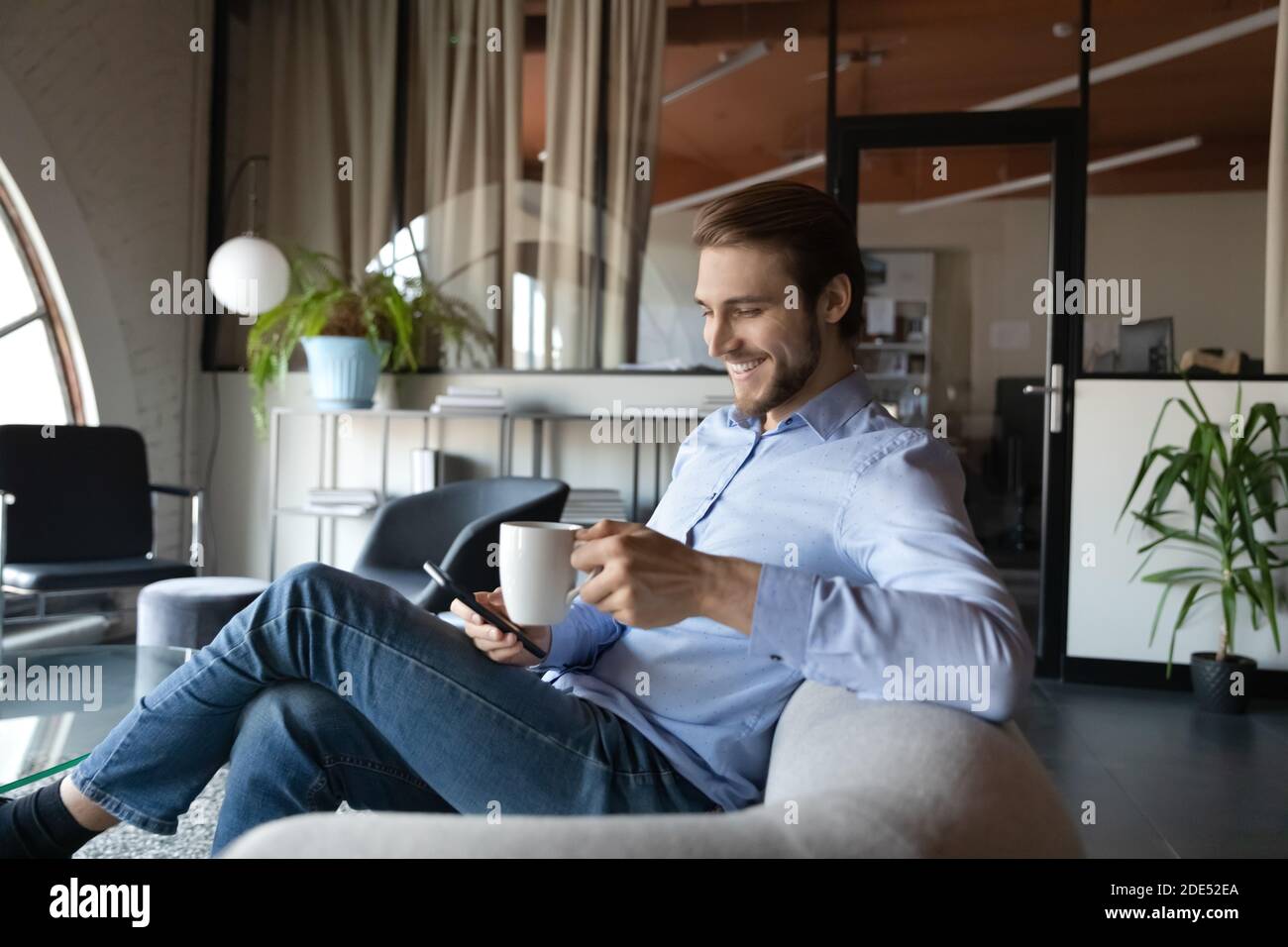Smiling man employee enjoying hot drink in office using cellphone Stock Photo