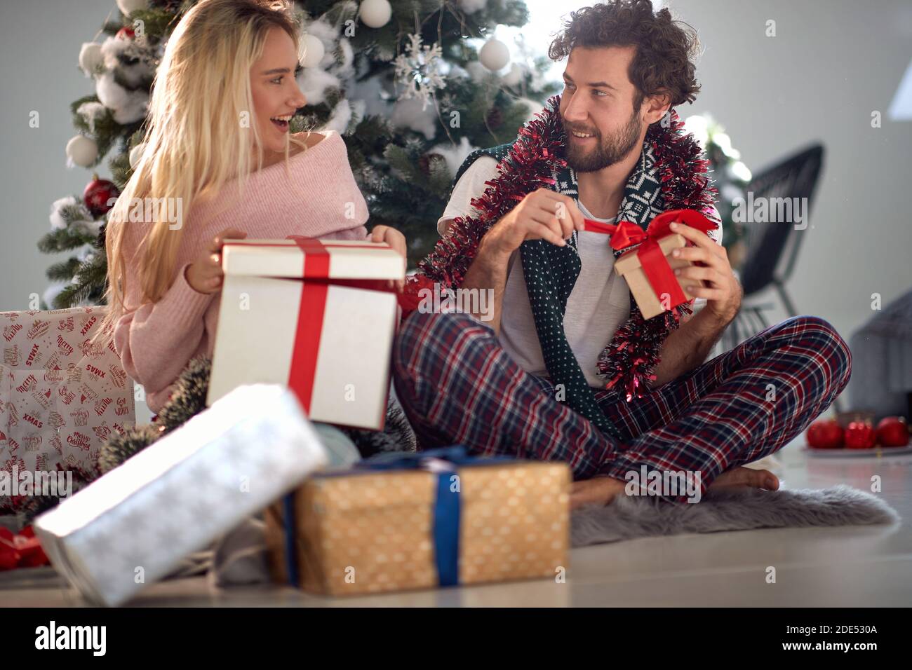 young Caucasian couple opening Christmas presents, smiling, looking each other, sitting on the floor in pajamas Stock Photo