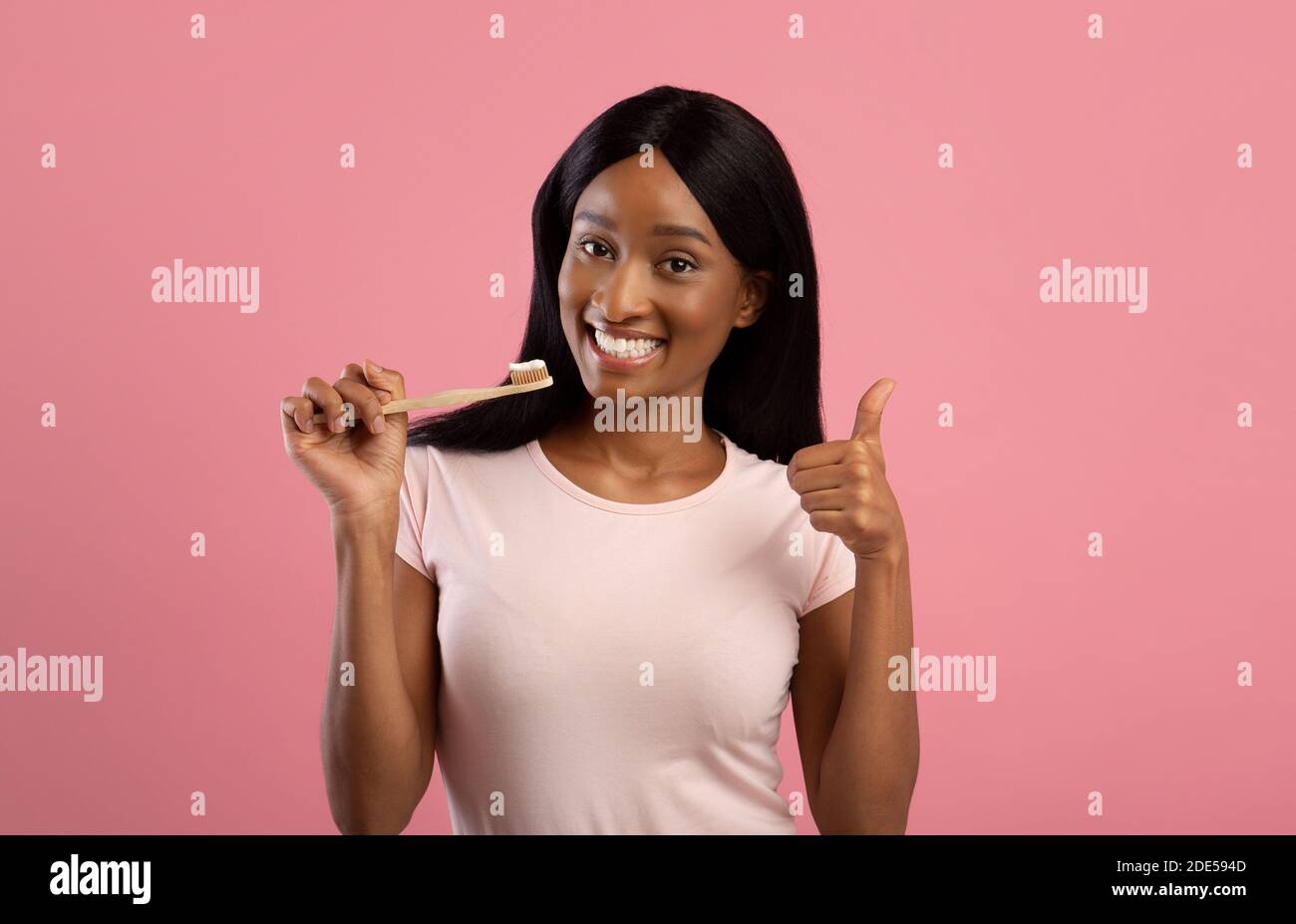 Pretty black lady with beautiful smile holding wooden toothbrush and showing thumb up gesture on pink background Stock Photo