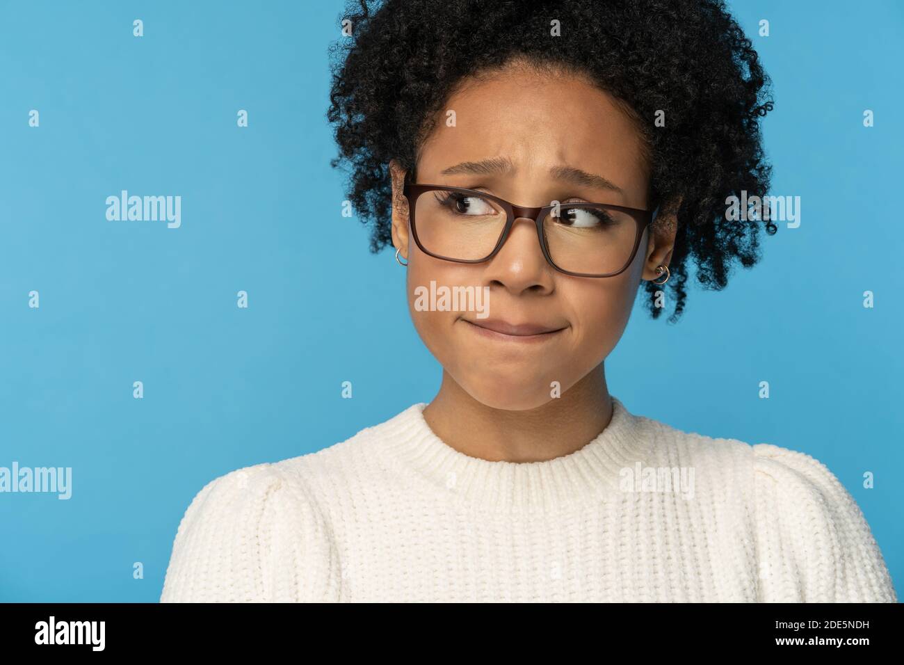 Close up studio portrait of shy awkward young Afro American woman wear glasses white sweater, biting lips feeling embarrassed, confused and nervous, l Stock Photo