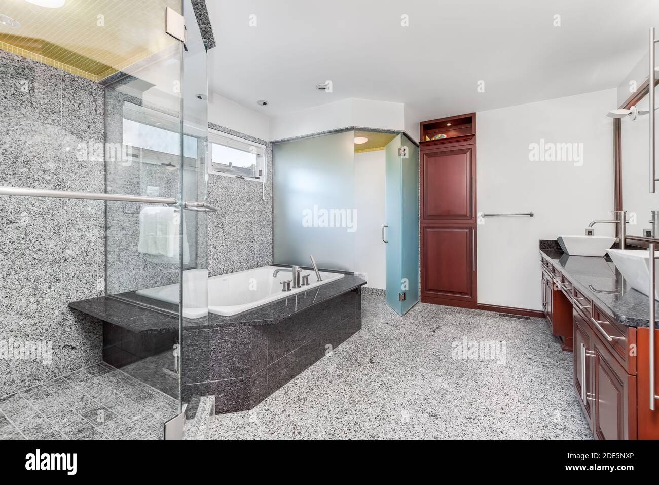 A large, modern bathroom with grey and white marble tiles on the floor and wall with a glass shower, jacuzzi tub and wood cabinets. Stock Photo