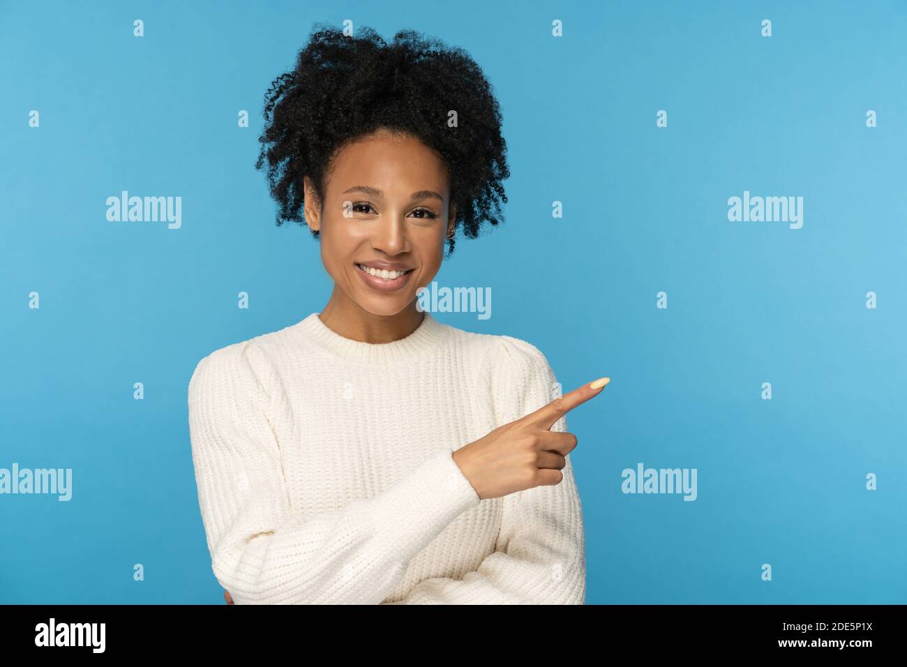 Smiling dark skinned woman with curly hair wear white jumper pointing with finger, showing blank copy space for advertising, offering, product, promot Stock Photo