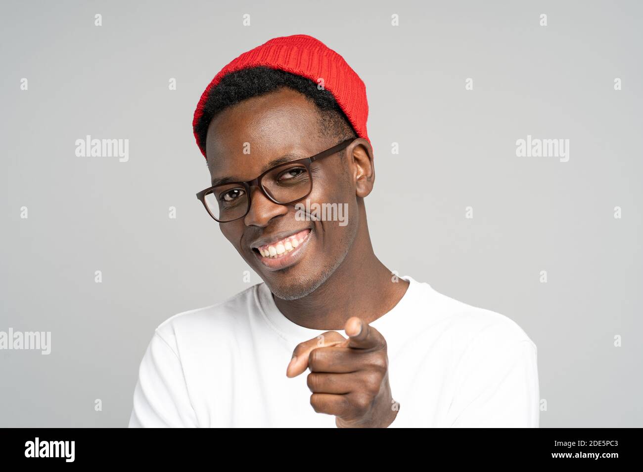 Hey, You! Cheerful positive young Afro American man wear red hat in good mood smiling broadly, pointing you while standing against studio grey backgro Stock Photo