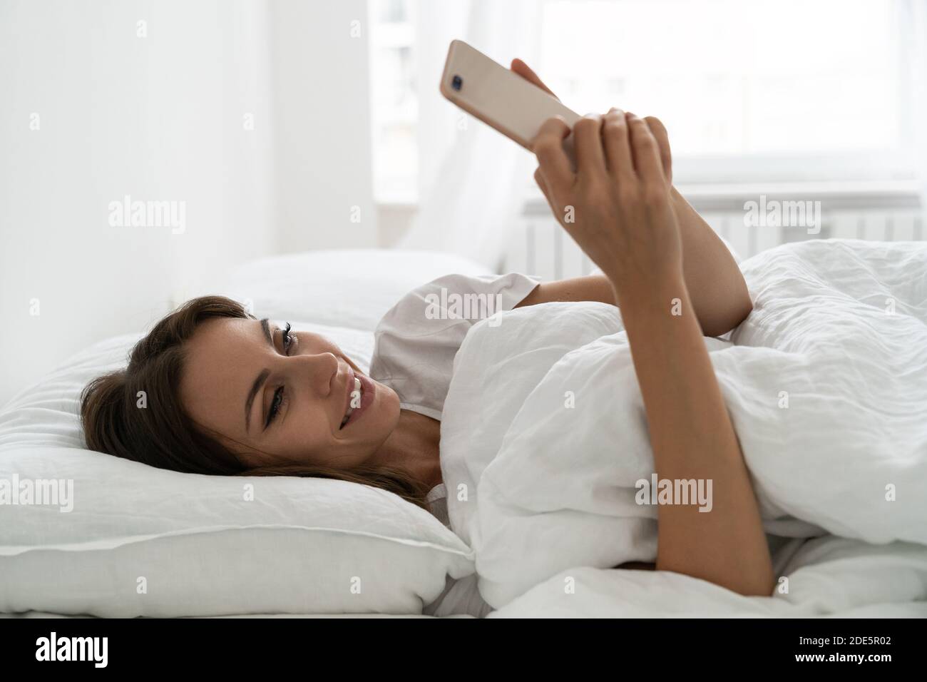 Female enjoying lazy weekend morning. Smiling millennial young woman using mobile smart phone, lying and relaxing in white comfortable bed at home, ch Stock Photo