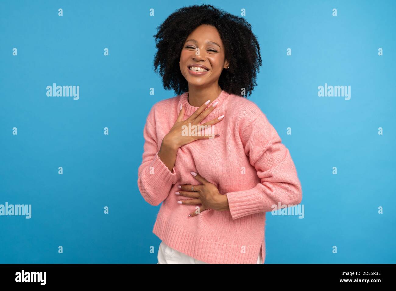 Laughing mixed race millennial woman with curly hair in casual pink jumper, keeps hand on chest, having fun, isolated on studio blue background. Overj Stock Photo