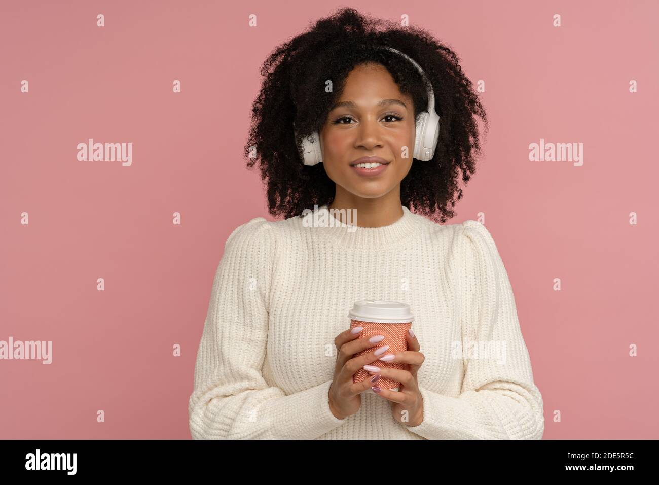 Cheerful positive dark skinned millennial woman with headphones wear white sweater looking at camera, holding paper cup of hot coffee or tea takeaway. Stock Photo