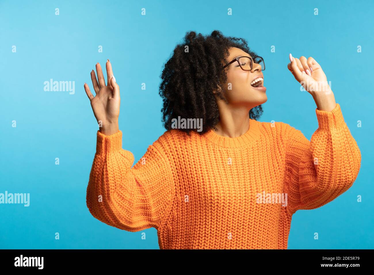 Funny dark skinned millennial woman with curly hair in good mood sings her favorite song, dancing, wear hipster oversize orange sweater, isolated on s Stock Photo