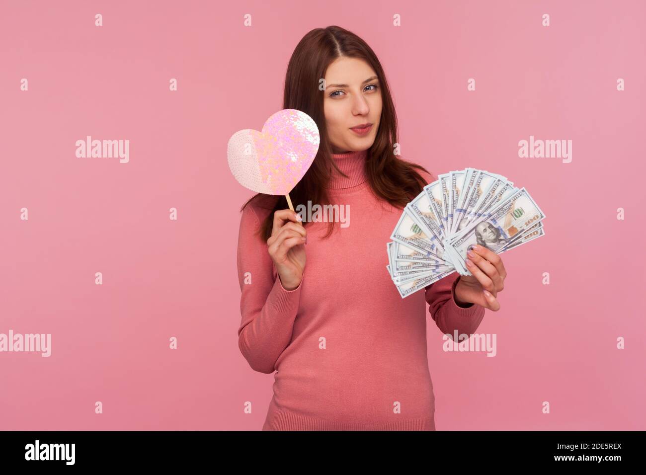 Cunning mercantile brunette woman in pink sweater holding paper heart and dollar banknotes and smiling looking at camera. Indoor studio shot isolated Stock Photo