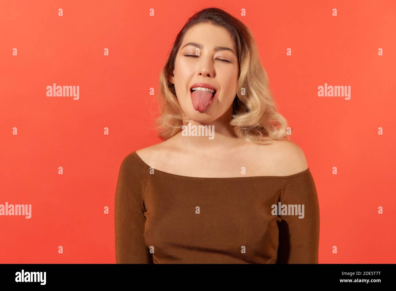Portrait of funny disobedient young woman with blonde hair keeping eyes closed and demonstrating tongue, behaving naughty unruly, childish mood. Indoo Stock Photo