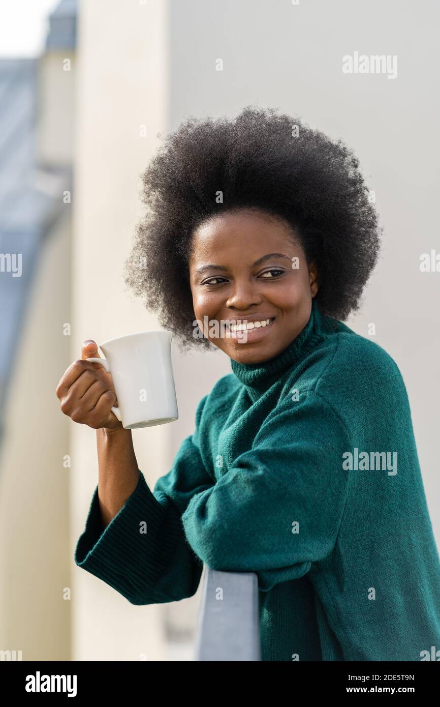 Smiling young African American biracial woman enjoying a cup of tea or coffee, wear oversize green knitted sweater, looking away, standing on the balc Stock Photo