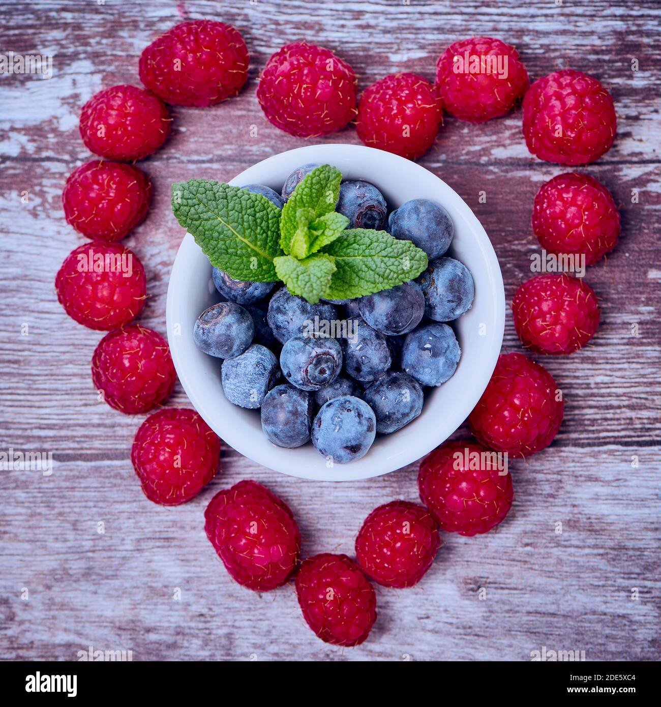 raspberries on wooden surface in a heart shape, there are blueberries in a white bowl in the middle of the heart, square color photo Stock Photo