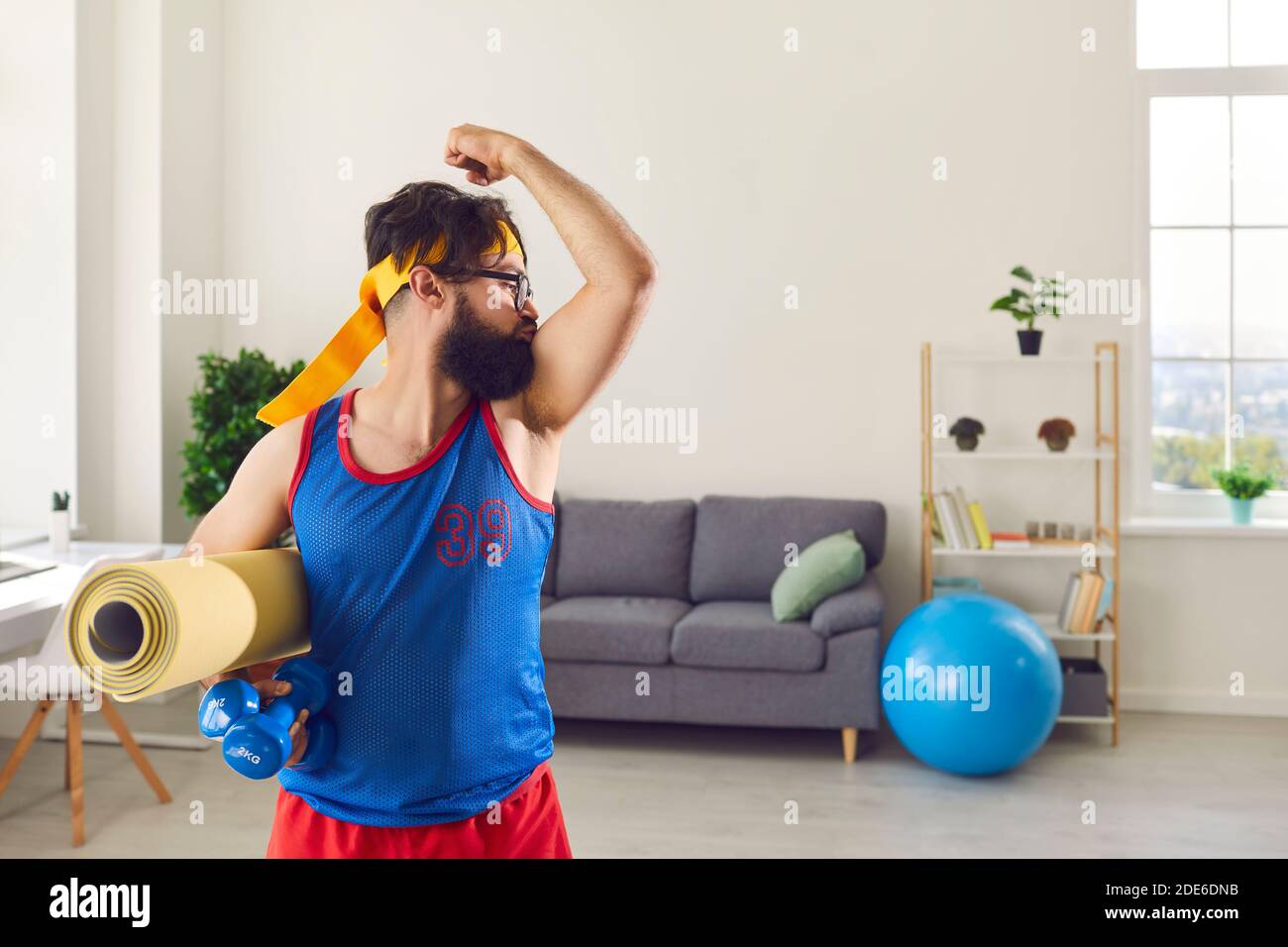 Funny bearded athlete in bright clothes and with a mat in his hands kisses his biceps. Stock Photo