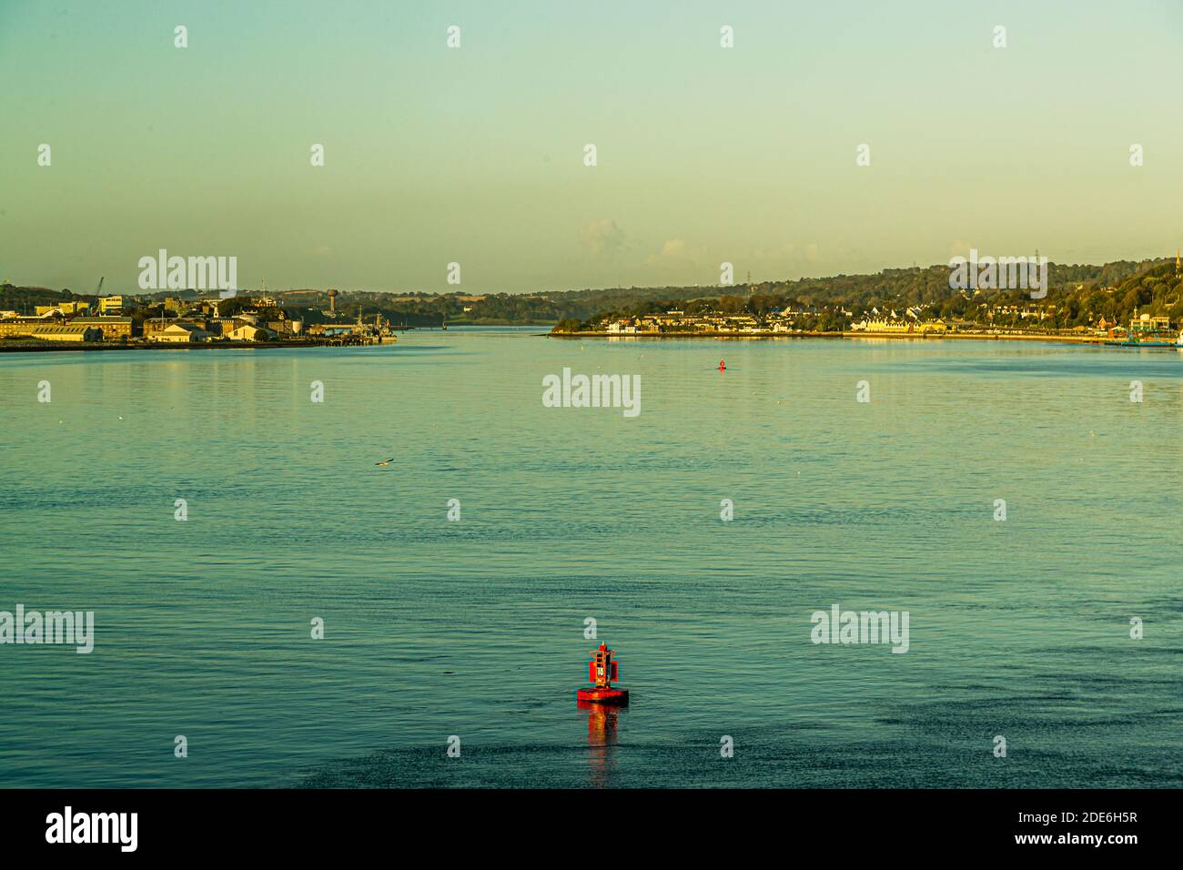 A red channel buoy marks the fairway in the harbour of Cork, Ireland, with settlements on both shores in the morning light. Stock Photo