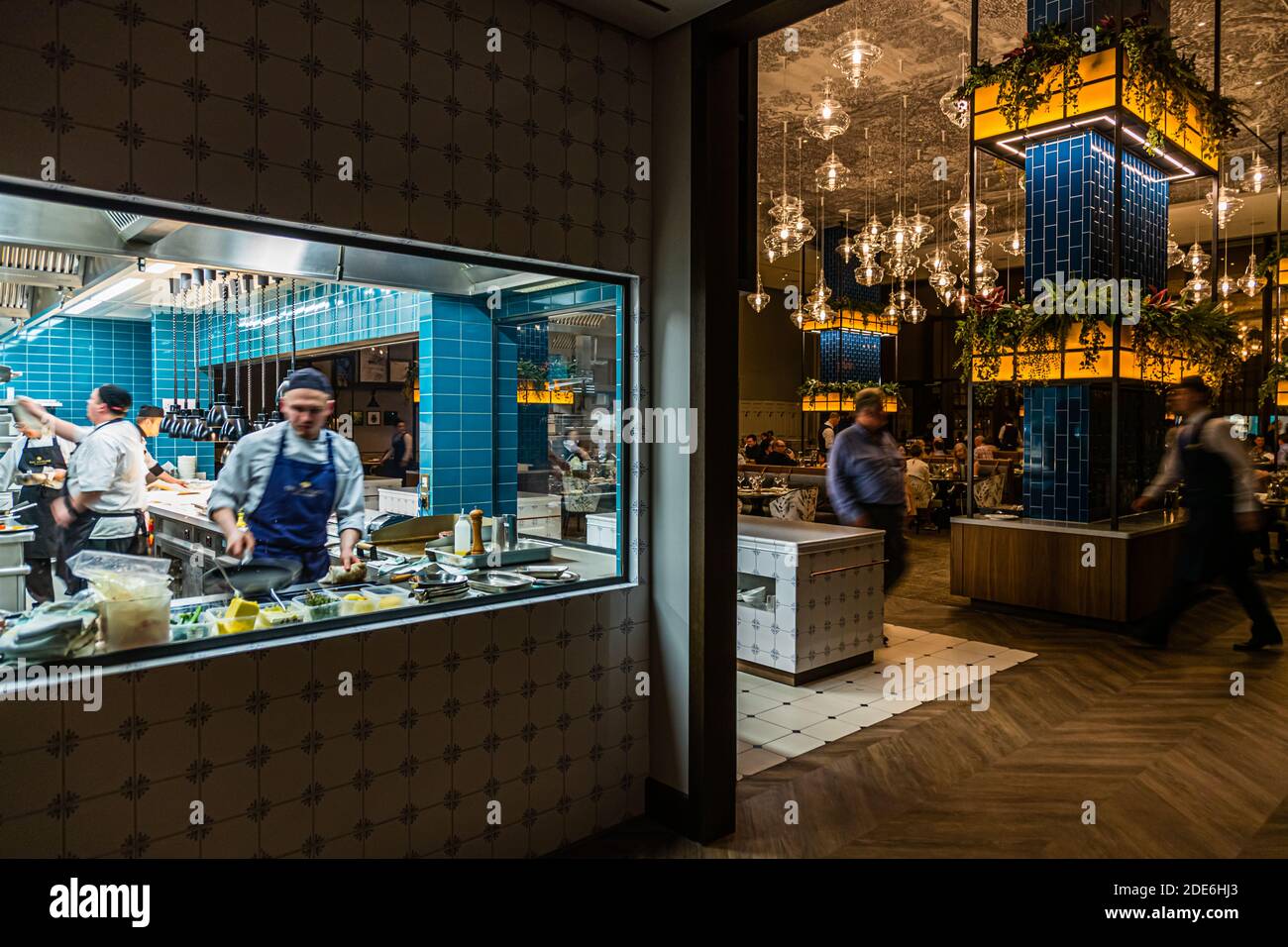 An open kitchen window connects the kitchen with the dining room of The Grill restaurant at the Dunloe Hotel & Gardens near Killarney, Ireland. The dining room features a blue-tiled pillar hung with plants and glass lamps. Stock Photo