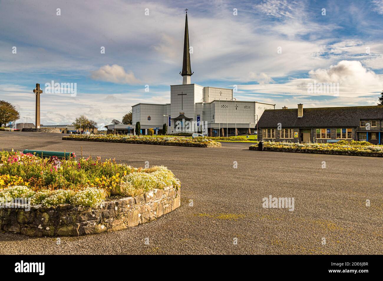 A Celtic stone cross stands before the Basilica of Our Lady, Queen of Ireland, on the grounds of the Knock Shrine Marian pilgrimage site in County Mayo, Ireland. Flower beds line the sprawling forecourt. Stock Photo