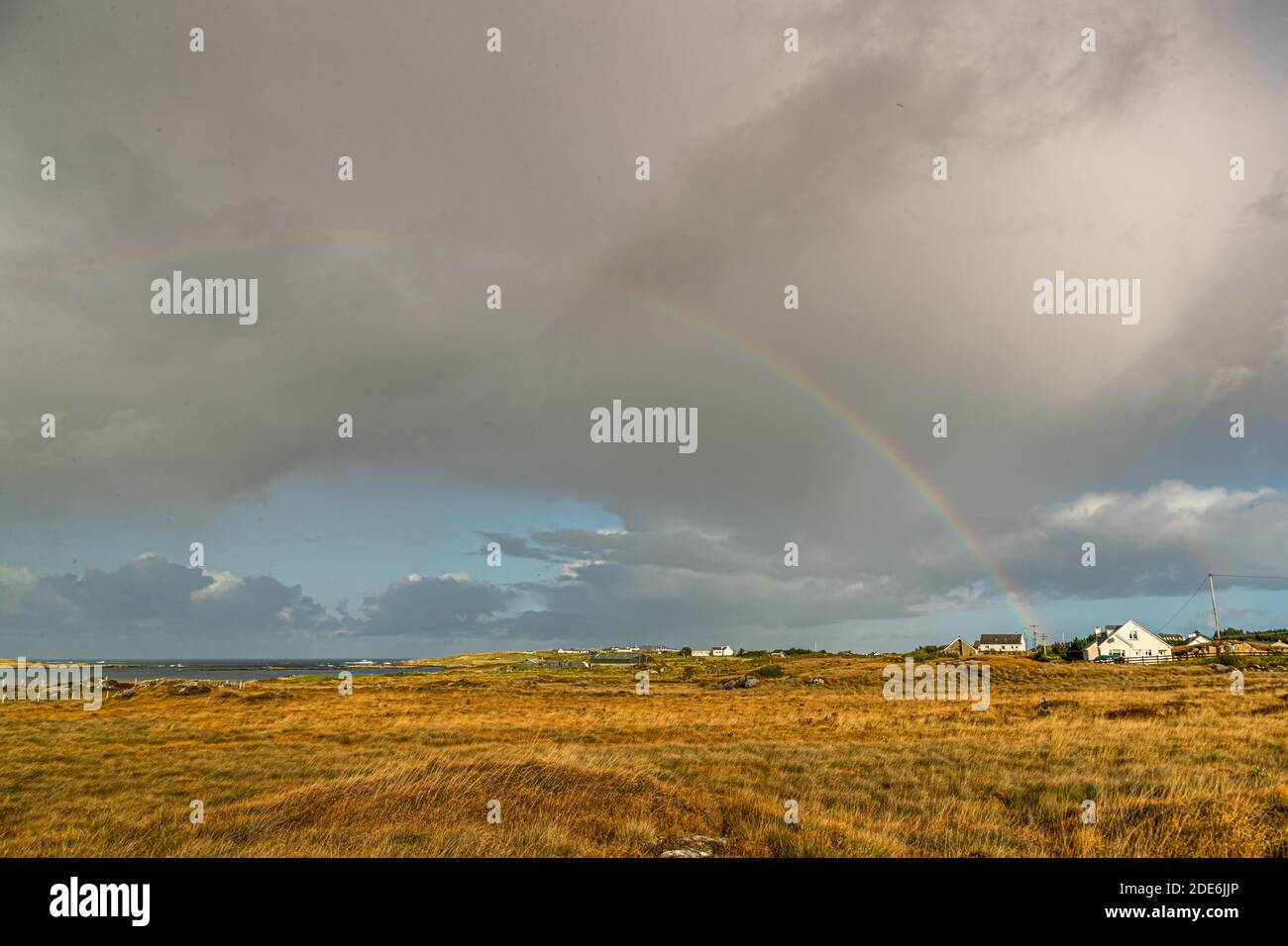 A rainbow glows over green hills along the R259 road in County Donegal, Ireland, beneath a cloud-streaked sky. Stock Photo