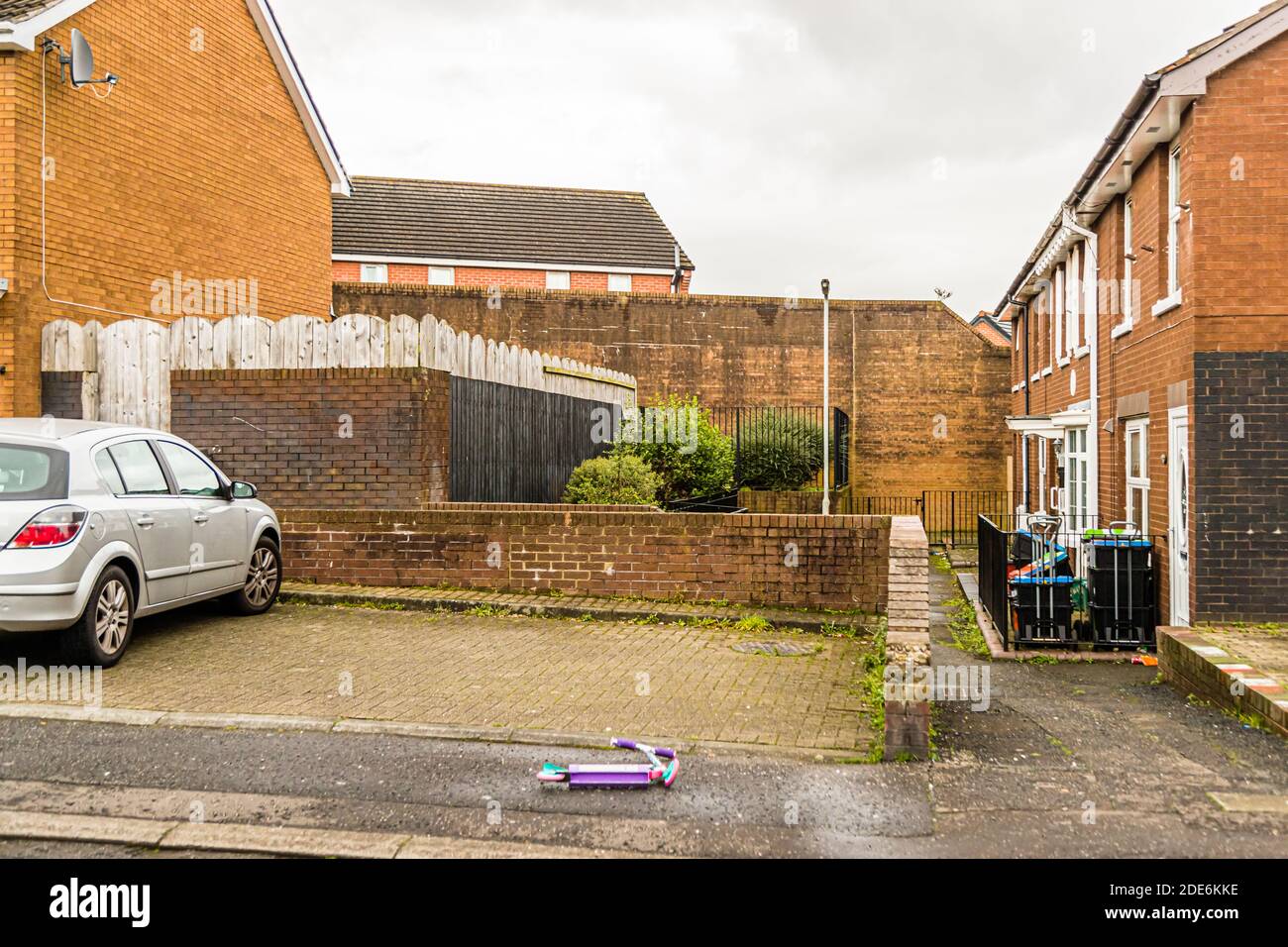 A quiet residential street in Clonard Rise, Belfast, Northern Ireland, ends at a tall peace-lines wall shielding the houses from a road beyond it; a child's scooter lies on the road. Stock Photo