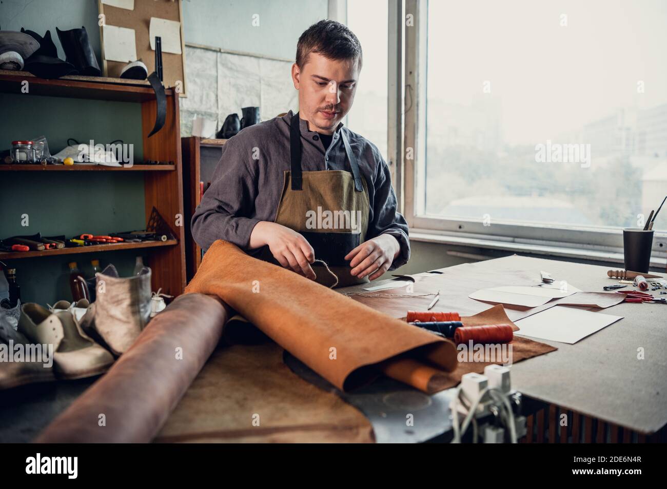 A young apprentice in a boot workshop prepares leather for further use on a large table. Stock Photo