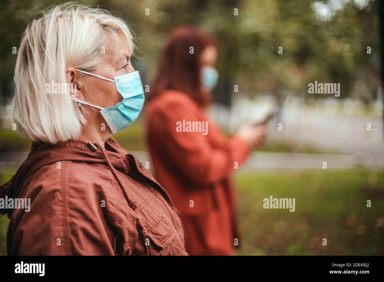 Coronavirus, Covid 19 epidemic. People wearing face mask outdoors in the autumn park. Girl uses a smartphone Stock Photo