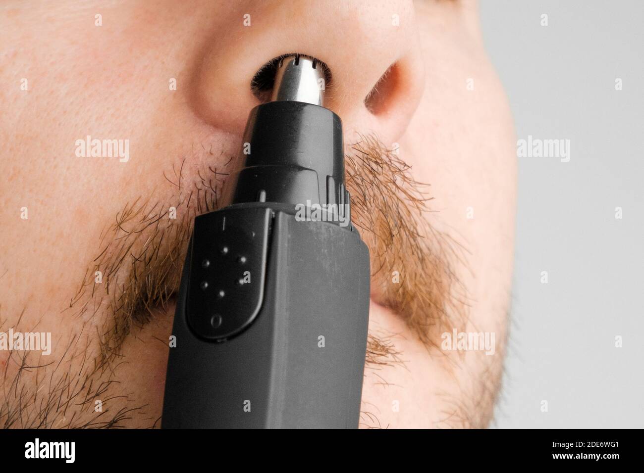 Close up man removing hair from nose using electric trimmer on the grey background. Morning routine. Stock Photo