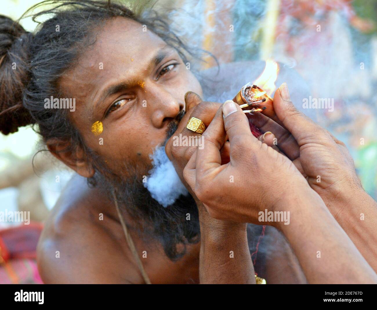 Hashish-smoking young happy Indian sadhu with top bun lightens his chillum pipe with the help of another sadhu during Bhavnath Fair (Shivratri Mela). Stock Photo