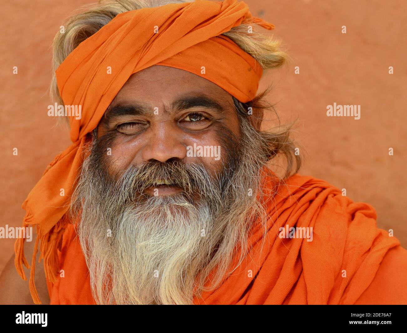 One-eyed elderly Indian Hindu holy man (sadhu, baba) with full beard, clad in orange, poses for the camera in front of a brown wall. Stock Photo