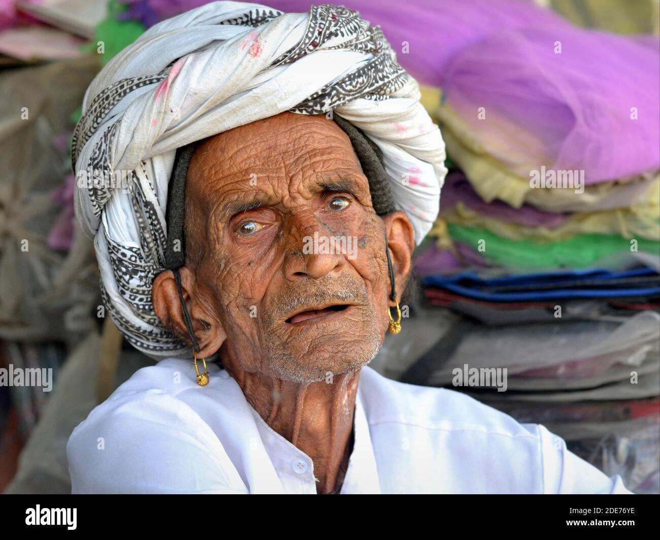 Frail old toothless Indian Rabari man with golden tribal earrings and wrinkled face wears a traditional turban and looks at the viewer. Stock Photo