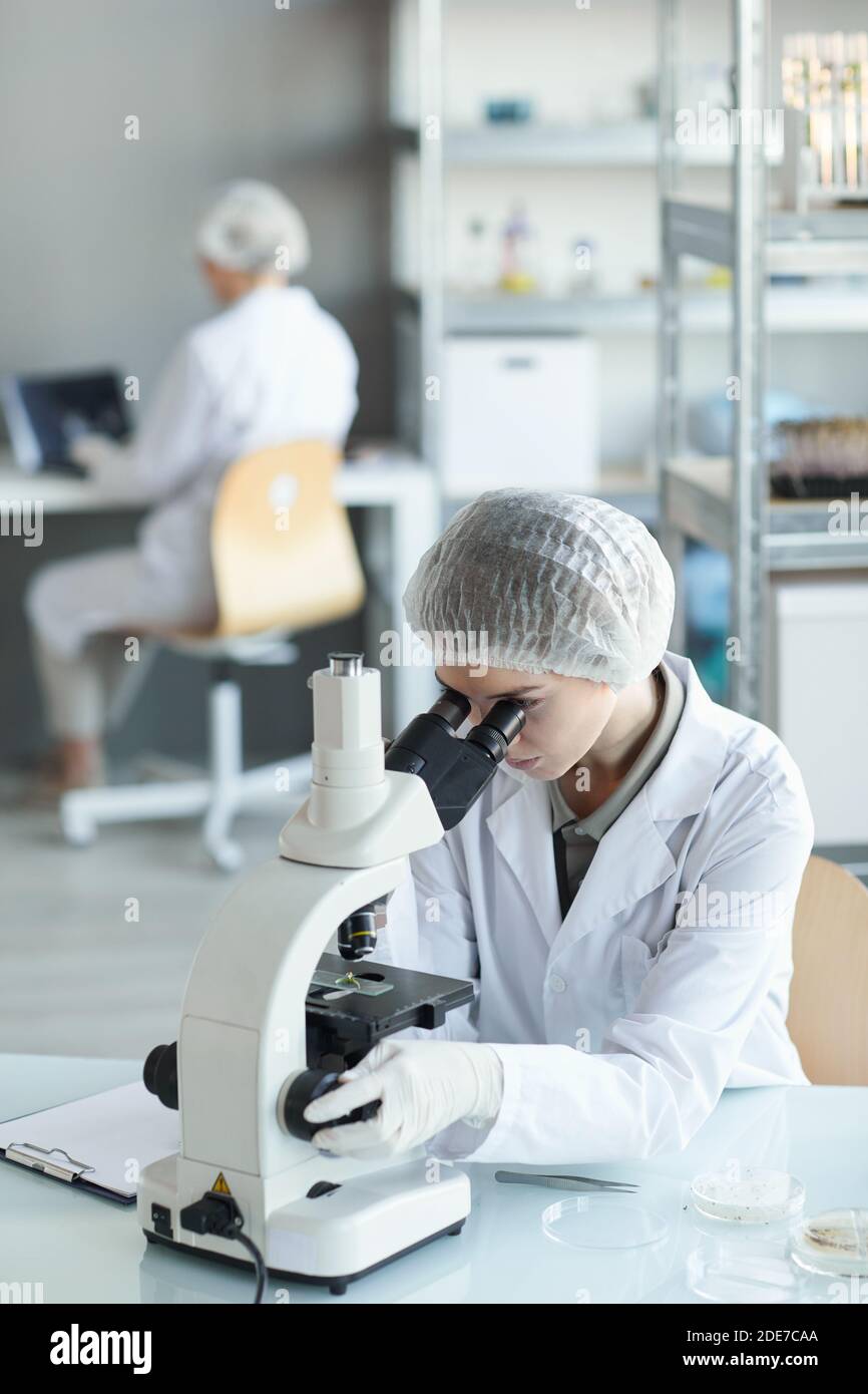 Vertical portrait of young female scientist looking in microscope while studying plant samples in biotechnology lab Stock Photo