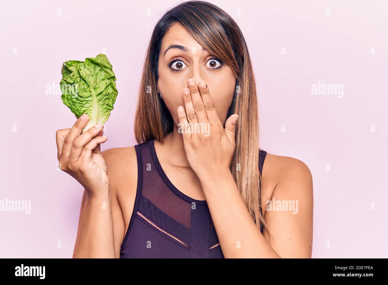 Young beautiful woman holding lettuce covering mouth with hand, shocked