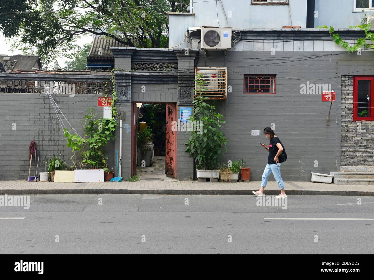 A woman checks her phone as she walks past the door of a Siheyuan courtyard house on a street in western Beijing, China Stock Photo