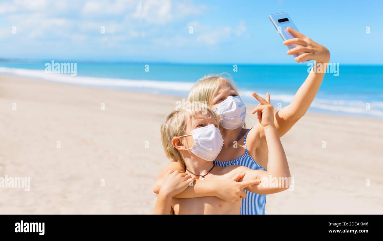 Funny kids taking selfie photo by smartphone on tropical sea beach. New rules to wear cloth face covering mask at public places due coronavirus COVID Stock Photo