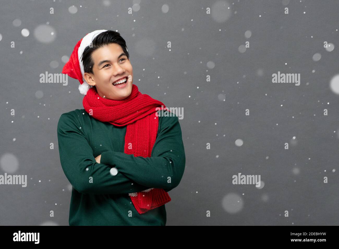 Smiling happy Asian man wearing colorful red and green Christmas theme clothes with arms crossed in snowfall on gray background Stock Photo