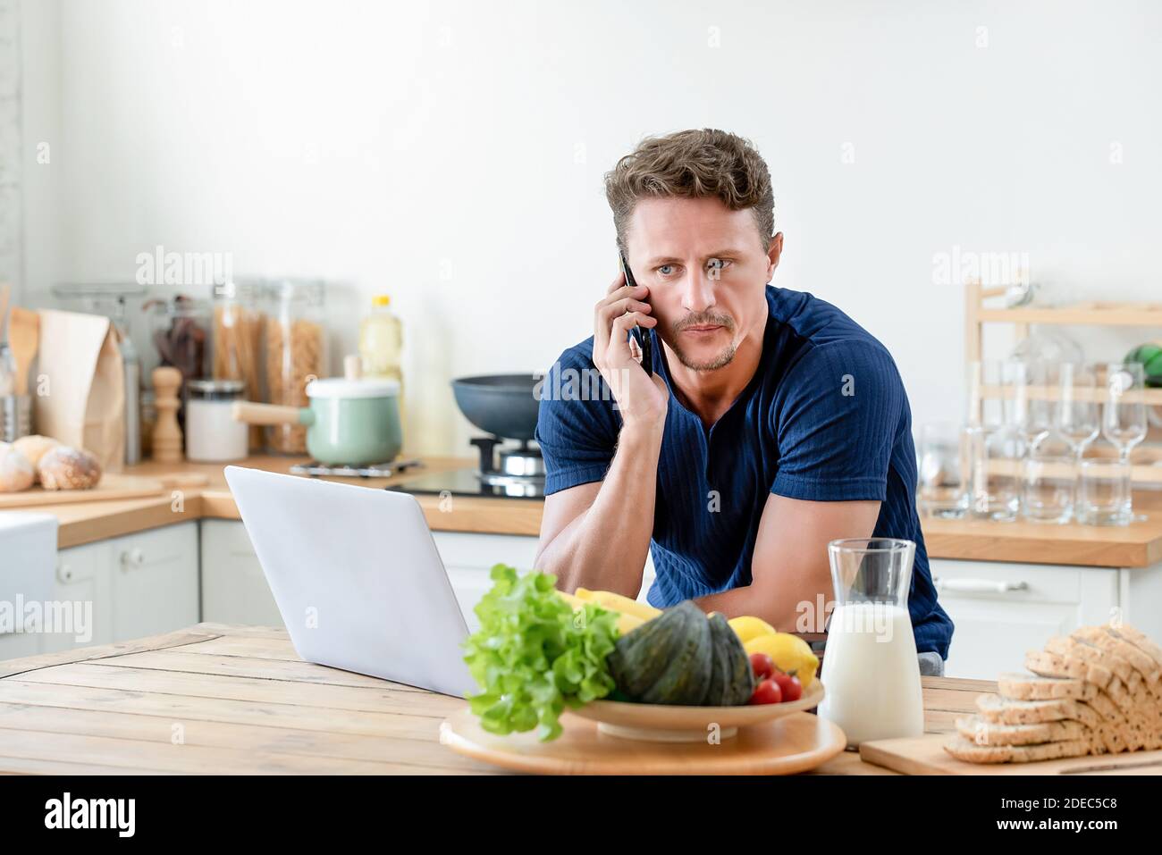 Casual man calling on cell phone while using laptop computer at home in the morning Stock Photo