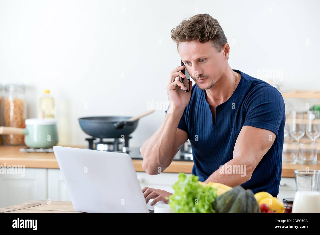 Casual man calling on cell phone while working on laptop computer at home in the morning Stock Photo
