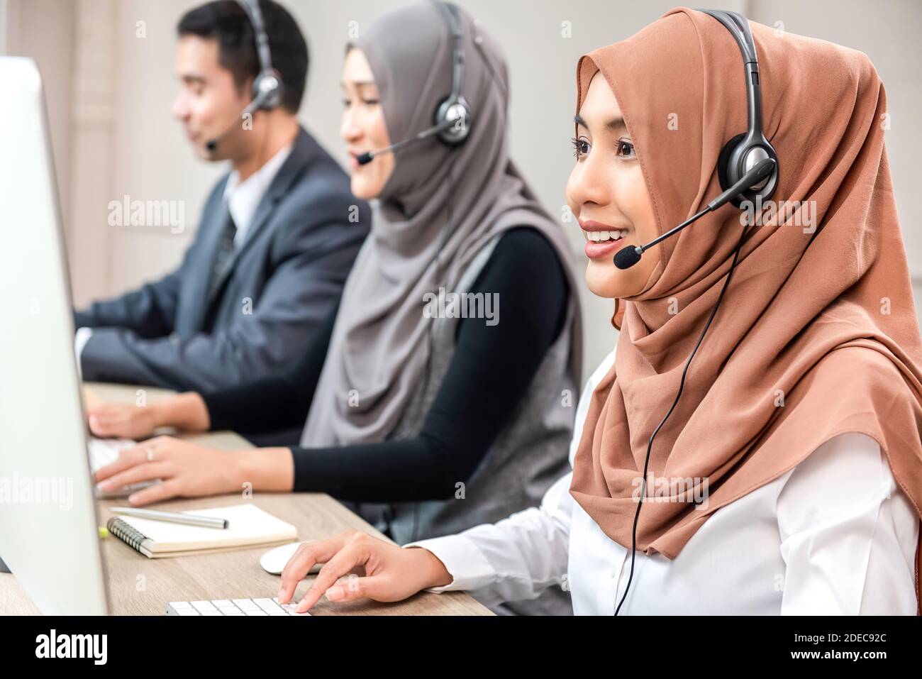 Friendly beautiful Asian muslim woman wearing microphone headset working as customer service operator with team in call center office Stock Photo