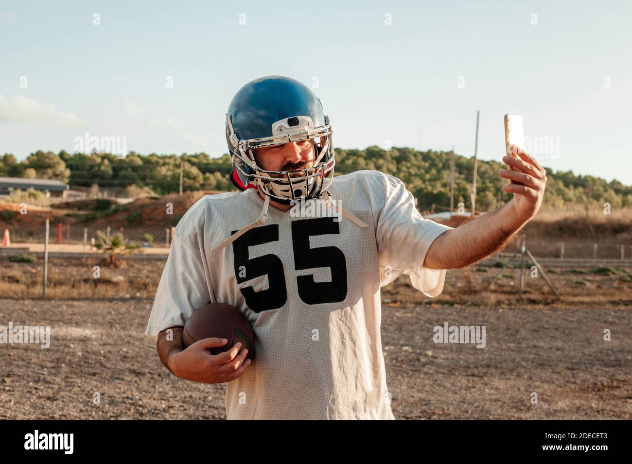 American football player using smartphone to take a selfie while holding a ball outdoors. Stock Photo