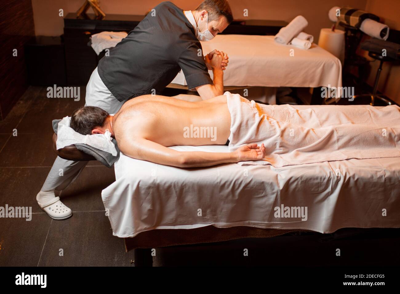 Professional masseur in facial mask doing a deep massage to a male client at Spa salon. Business during the epidemic concept Stock Photo