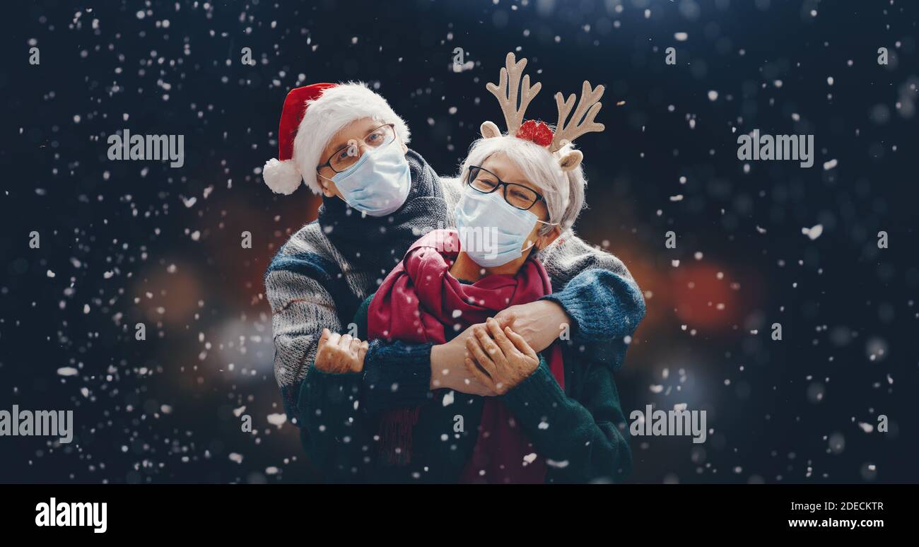 Merry Christmas! Winter portrait of senior couple on snowy dark background. People wearing facemasks. Stock Photo