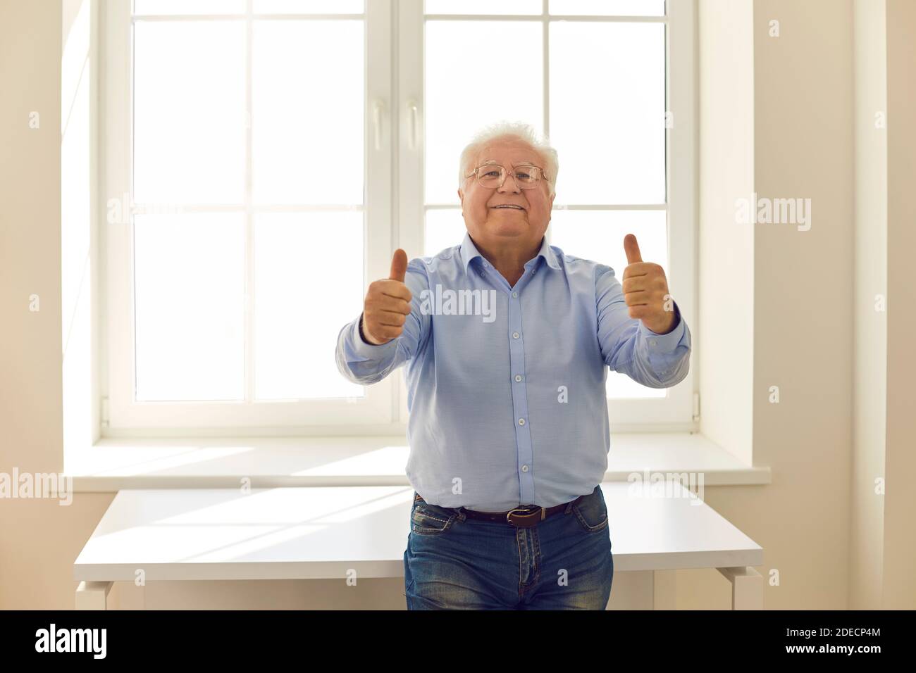 Positive senior man giving thumbs-up and looking at camera standing by the window at home Stock Photo