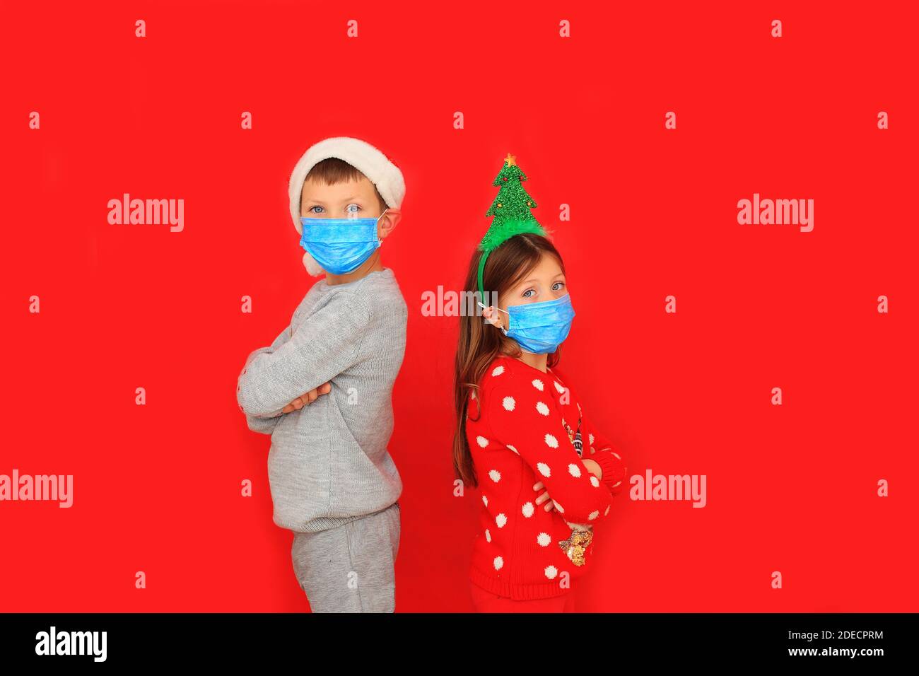 A boy and a girl wearing respiratory masks keep their distance on New Years during the quarantine period. Children in New Year's knitted sweaters on a red background Stock Photo