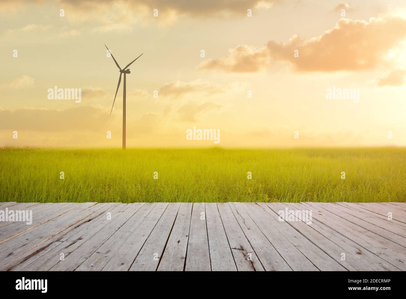 Wind turbine on green rice field against sunset background with plank wood foreground. Green energy concept. Stock Photo