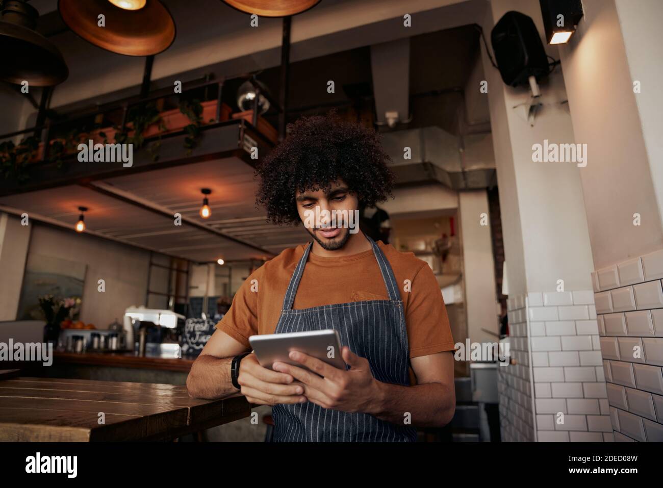 Young male waiter wearing apron uniform in cafe using digital tablet to watch recipe Stock Photo