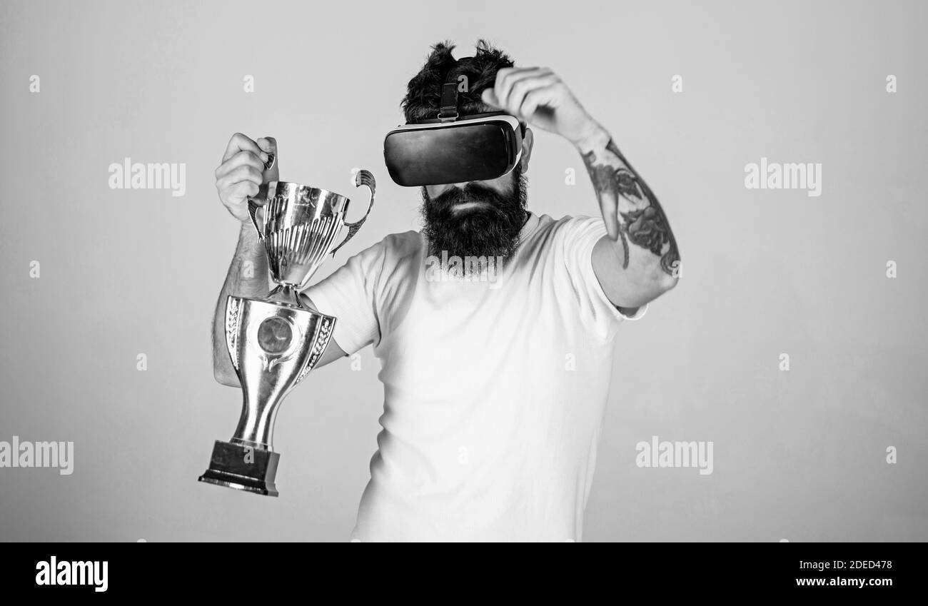 Guy with VR glasses won competition, holds in hand golden goblet. Man with  beard in VR glasses, grey background. Hipster on serious face use virtual  reality for achievements. Virtual victory concept Stock